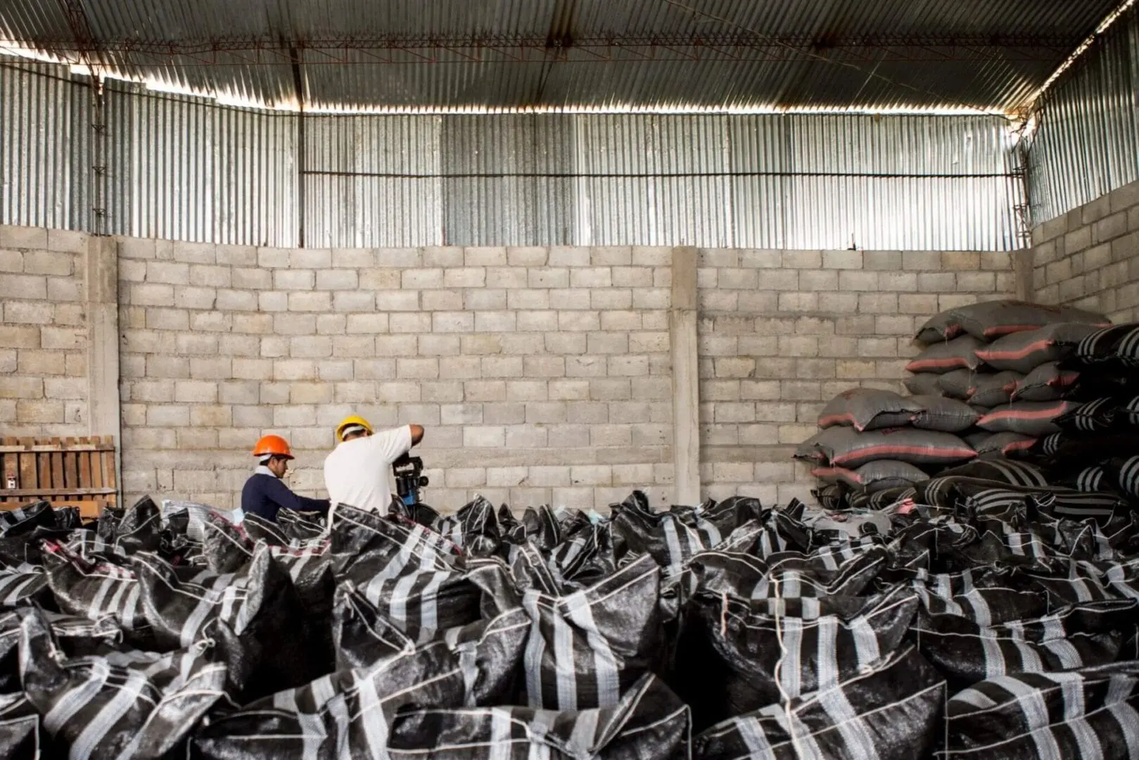 Two men in hard hats and helmets are sitting on a pile of bags.